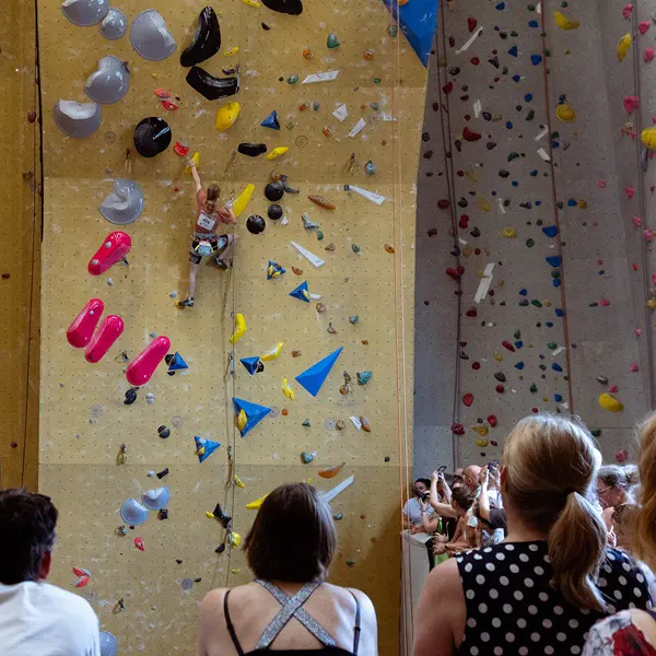 Een klimmer beklimt een indoor-klimwand met kleurrijke grepen, terwijl een menigte van beneden toekijkt. Sommige mensen op de voorgrond staan met hun rug naar de camera.